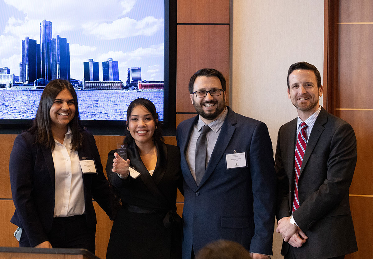 Four individuals (two women & two men) in business attire smile and pose for a photograph together next to each other as a woman from the group holds a chrome colored award plaque inside a conference room area at the Wayne State Jaffe/Taft Transactional Law Invitational in March 2023.