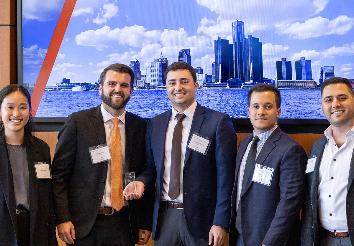 Five individuals (one woman & four men) in business attire smile and pose for a photograph together next to each other as a man from the group holds a chrome colored award plaque inside a conference room area at the Wayne State Jaffe/Taft Transactional Law Invitational in March 2023.