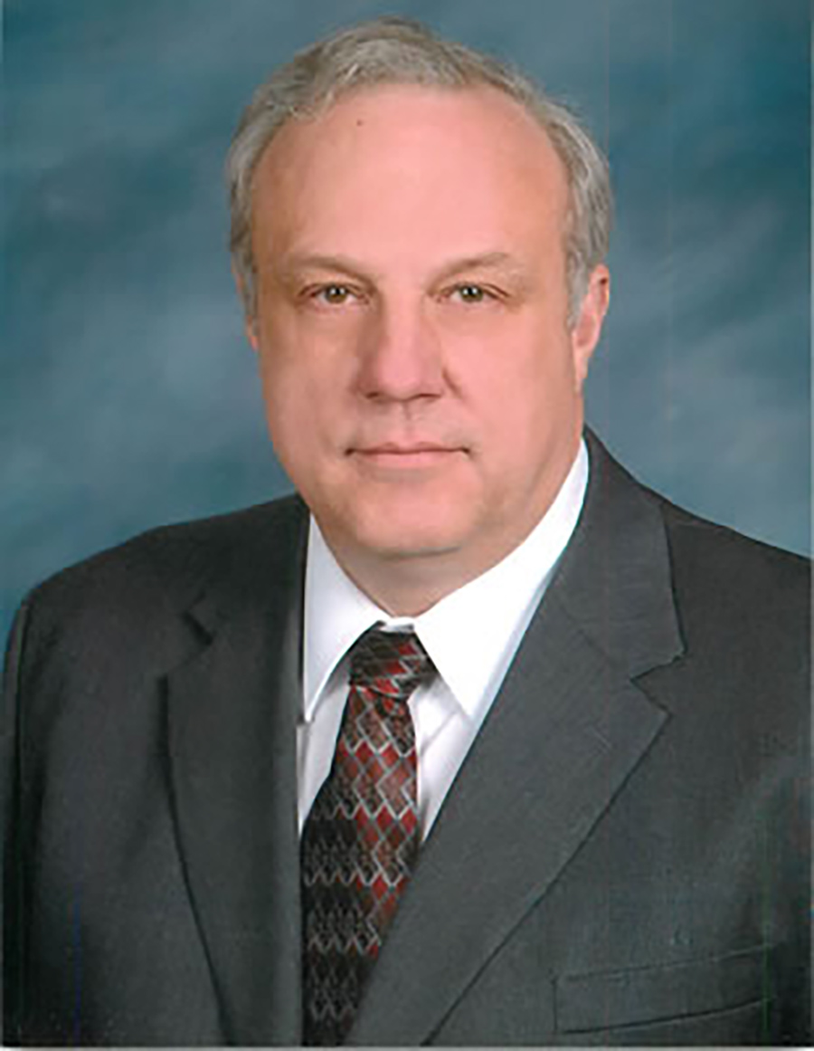 Portrait headshot photograph of John E. Walus grinning in a grey colored suit and white button-up dress shirt underneath with a multi-colored tie (red, black, white, and grey pattern style)