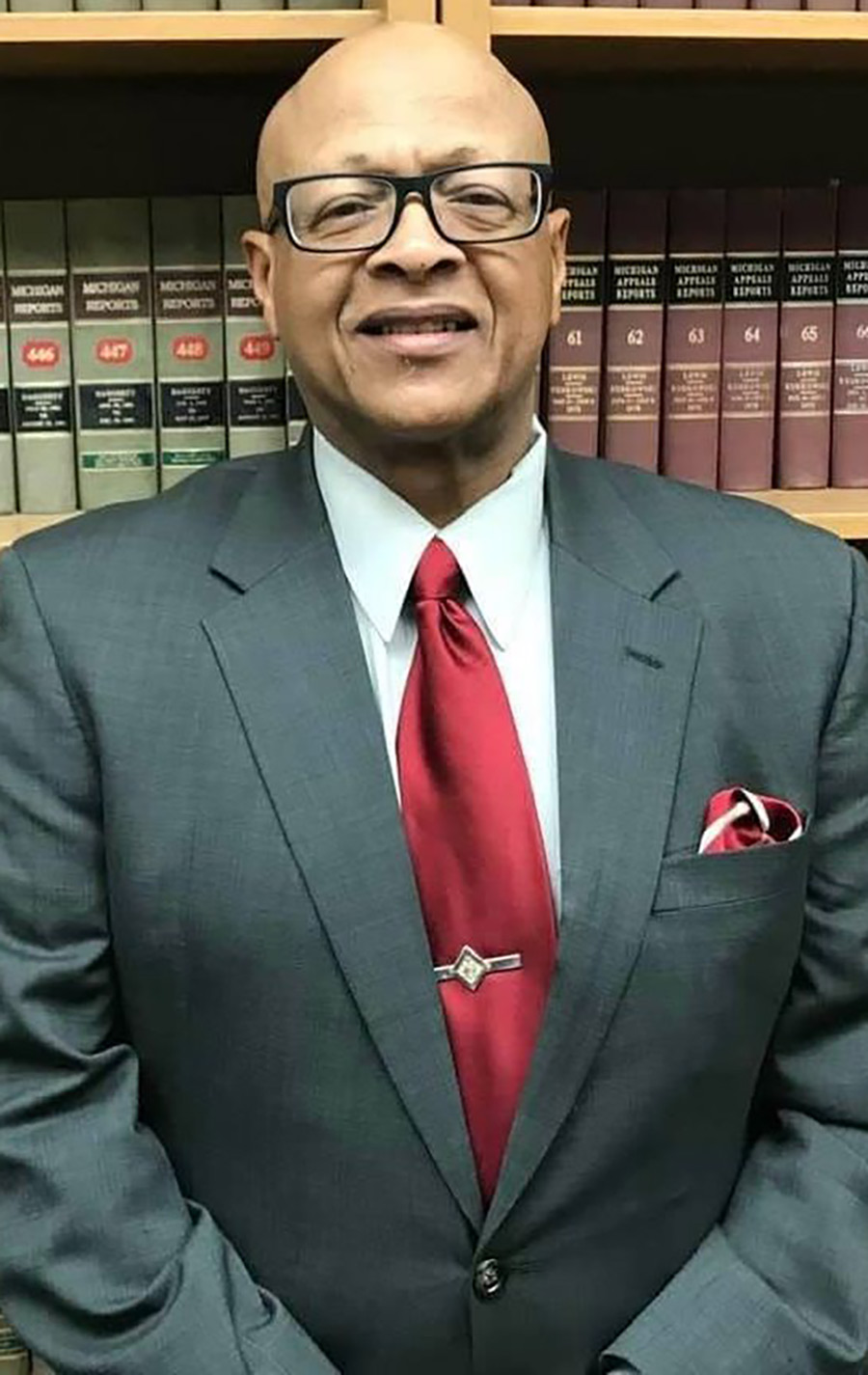 Portrait headshot photograph of Richard Smart III smiling in a grey colored suit and white button-up dress shirt underneath with a red colored tie in front of a bookshelf stack with his hands crossed for a pose