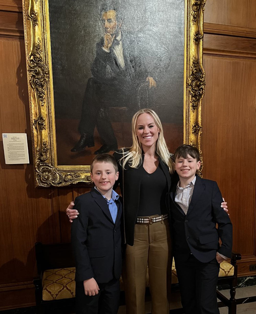 Shea Mace stands with an arm around each of her sons in front of a bench and a painting of Abraham Lincoln