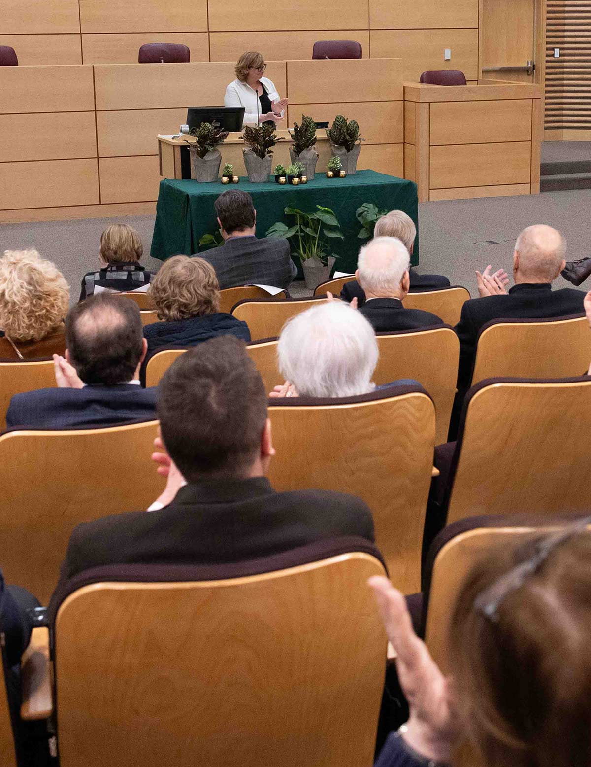 A woman in a white blazer gives a presentation at a podium in a tiered lecture hall. The audience is shown from behind in the foreground, with several people clapping.