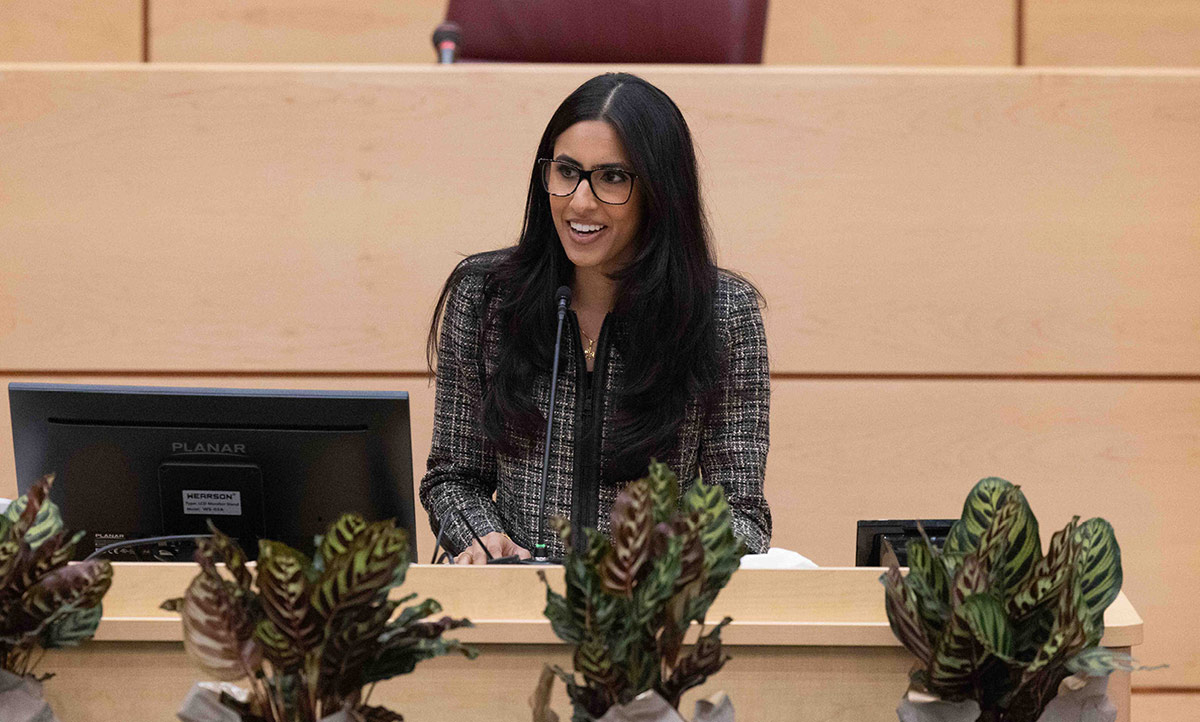 A woman with long dark hair and glasses speaks behind a podium in a lecture hall. She is wearing a grey tweed jacket, with potted plants visible in the foreground.