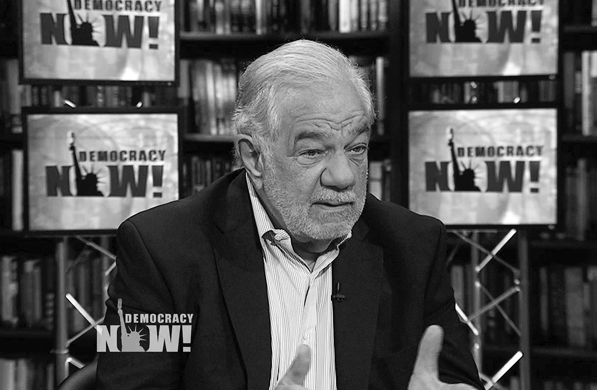 Black and white photo of an older man with a beard and suit jacket speaking on the set of "Democracy Now!". Several screens displaying the show's logo and a bookshelf are visible in the background.